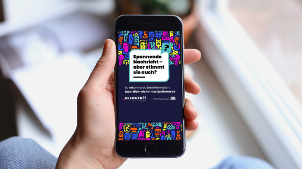 Smartphone mit GELOGEN?!-Kampagnenmotiv: Sprechblase „Spannende Nachricht – aber stimmt sie auch?“ und URL lass-dich-nicht-manipulieren.de
